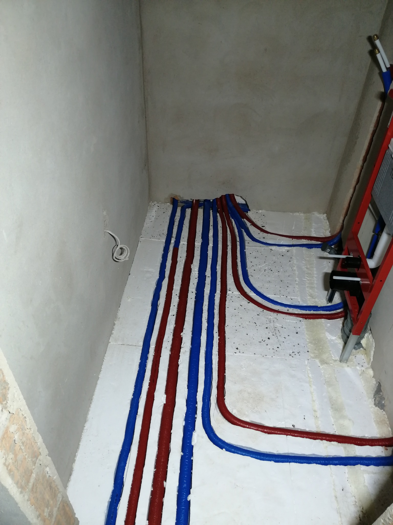 Montaż instalacji ogrzewania podłogowego z rurami w kolorach niebieskim i czerwonym, ułożonymi na białej izolacji, widok z góry w surowym wnętrzu łazienki.
