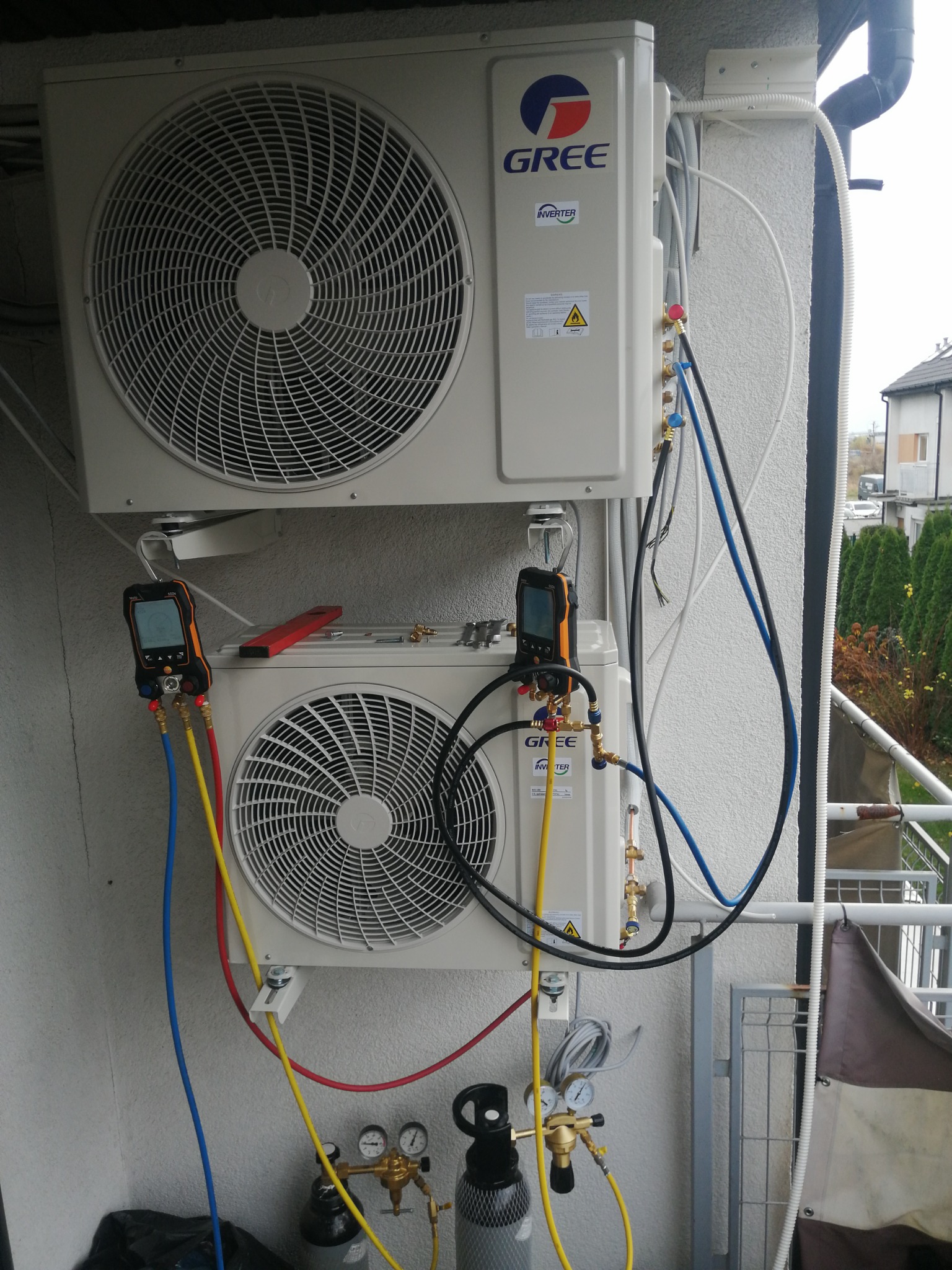 Dwa zamontowane klimatyzatory GREE na balkonie z podłączonymi narzędziami diagnostycznymi i butlami z gazem, widoczne przewody i manometry.