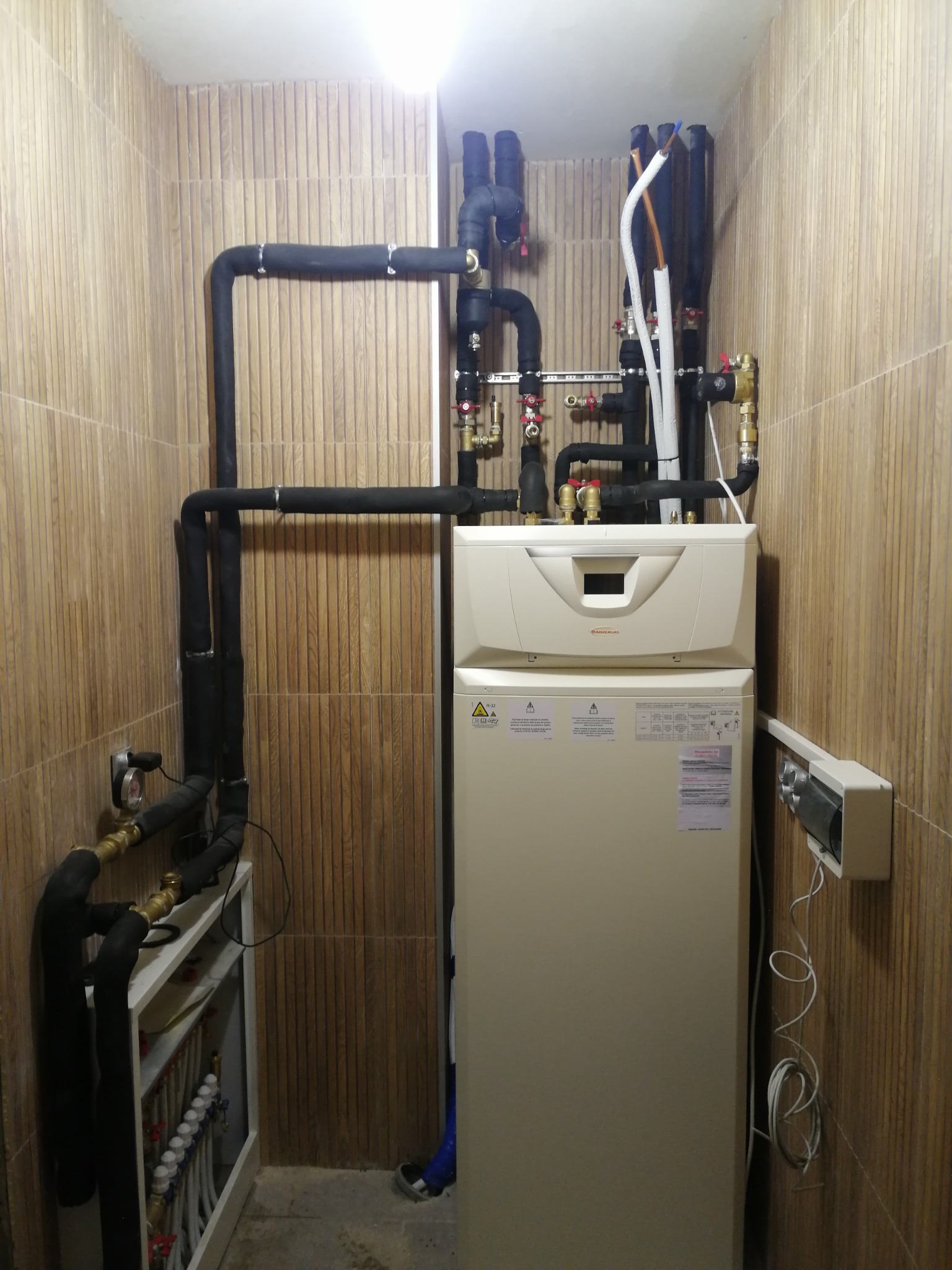 Zamknięte pomieszczenie z zamontowaną pompą ciepła Immergas, rurami w izolacji i rozdzielaczem ogrzewania podłogowego na tle ściany wyłożonej płytkami.