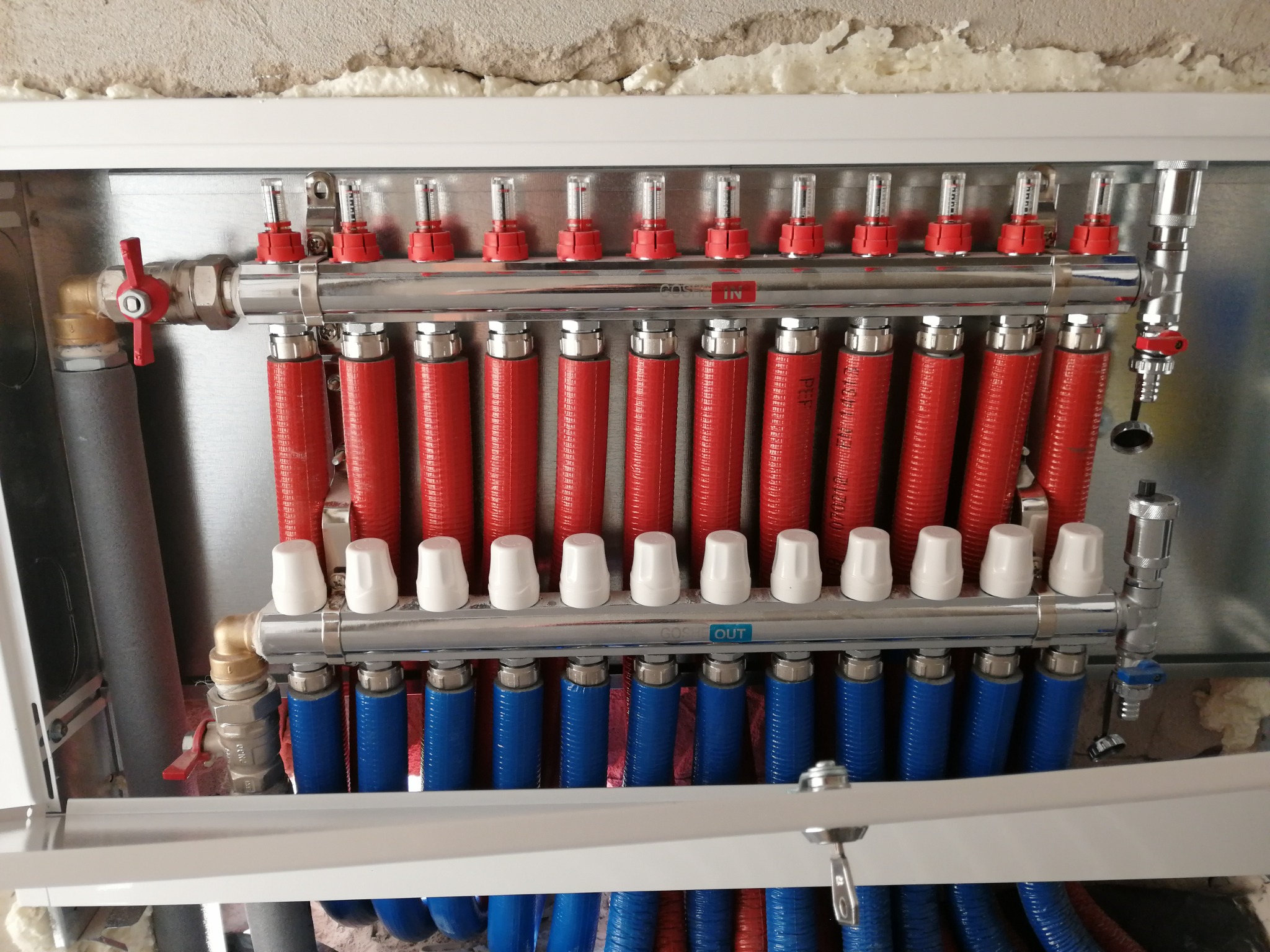 Rozdzielacz ogrzewania podłogowego z czerwonymi i niebieskimi rurami w metalowej szafce, widoczne zawory i mierniki przepływu. Instalacja w surowym wnętrzu.