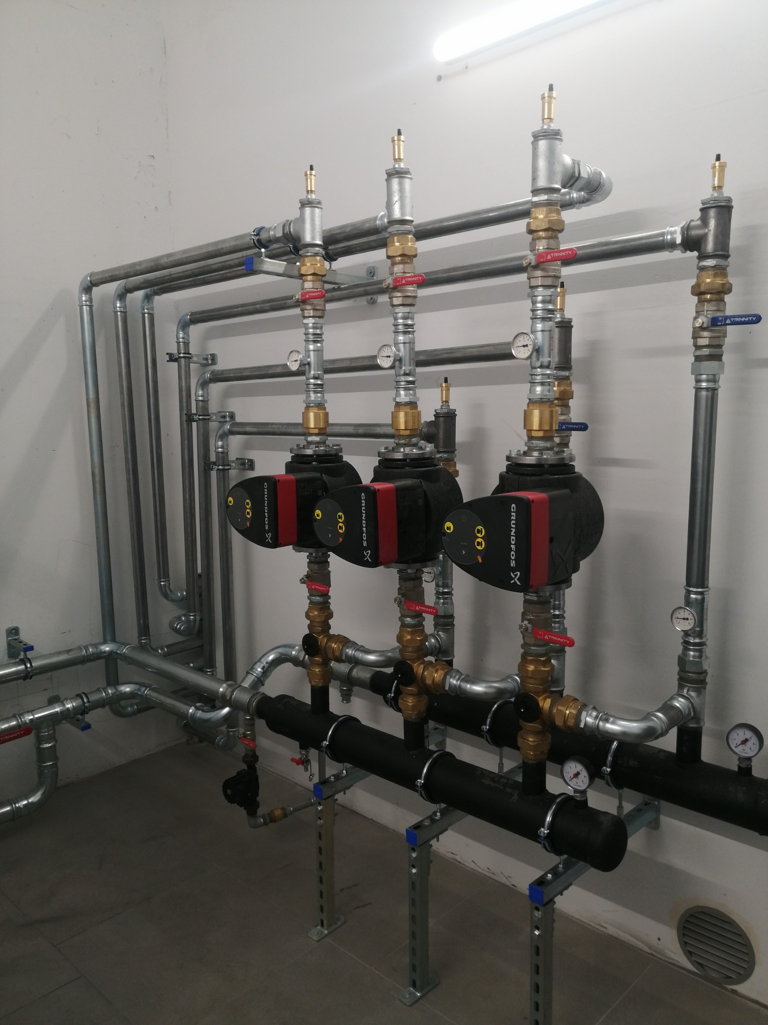 Złożona instalacja hydrauliczna z trzema pompami obiegowymi Grundfos, rurami stalowymi i mosiężnymi zaworami, zamontowana na wspornikach w pomieszczeniu technicznym.