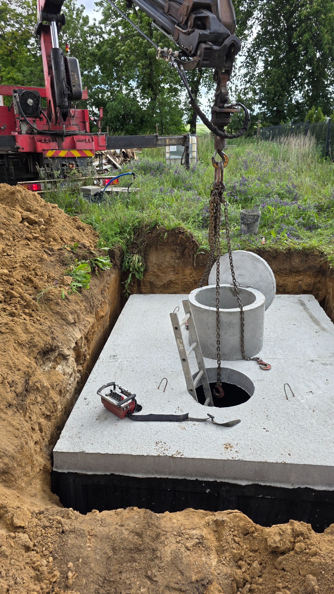 Montaż betonowego zbiornika: dźwig opuszcza krąg do wykopu z hydroizolacją, drabina oparta o płytę. Widoczny pilot sterujący i ziemia wokół.