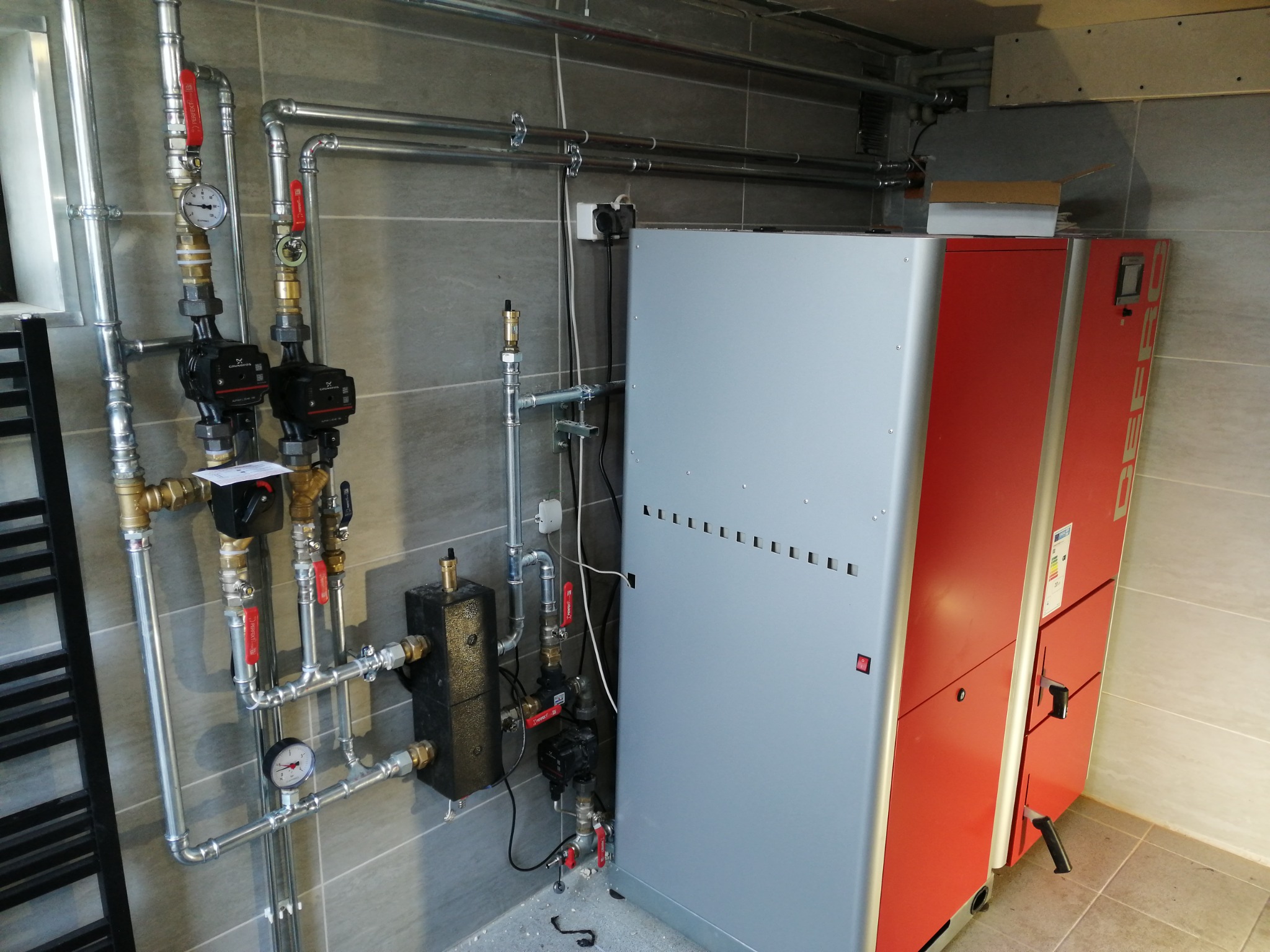 Nowoczesny piec CO z instalacją: srebrno-czerwony piec, rury, zawory, manometry na tle szarej ściany z płytek. Kompaktowa kotłownia w minimalistycznym stylu.
