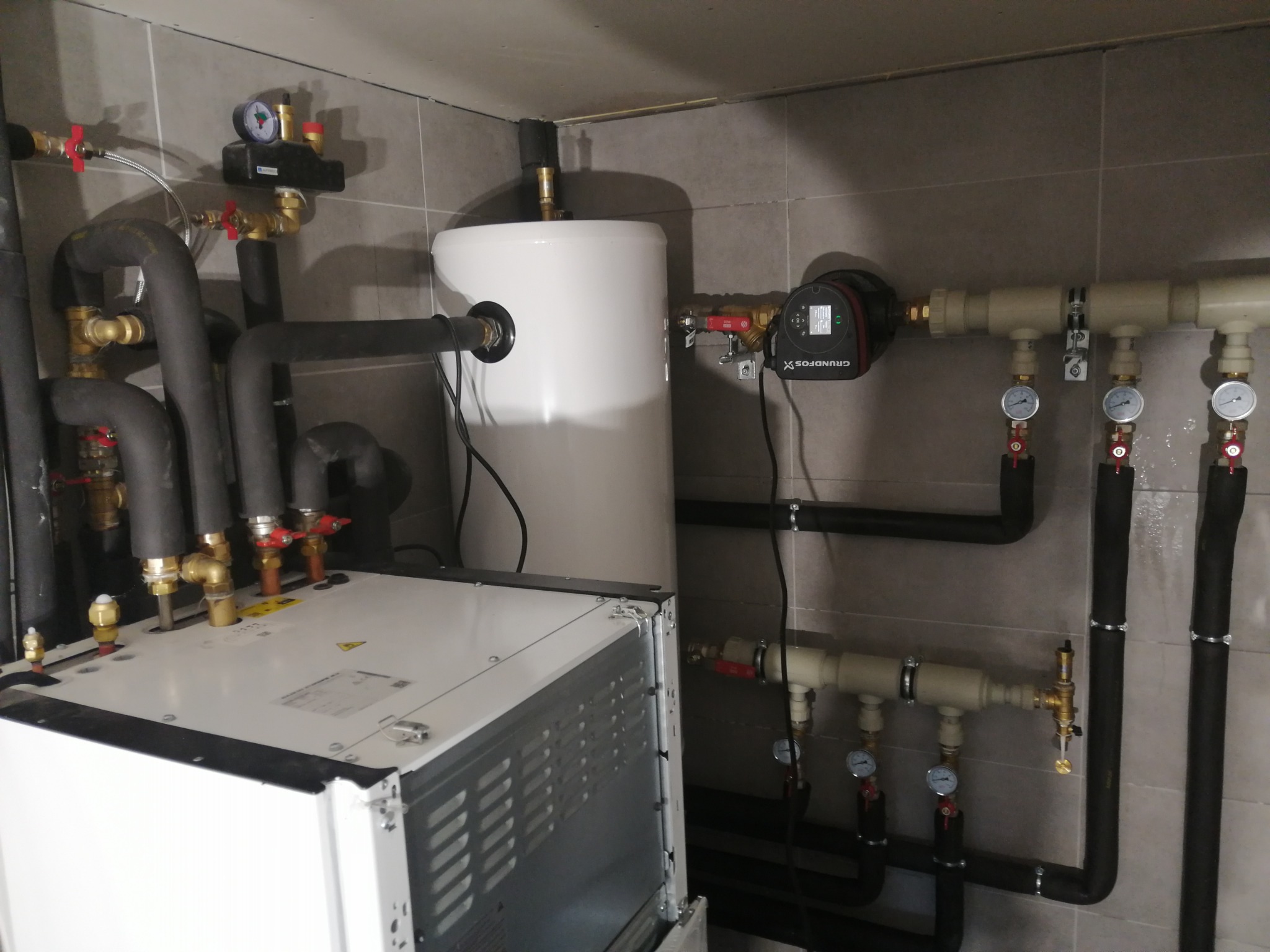 Kotłownia z białym zbiornikiem i czarnymi rurami, manometry i zawory. Widok na instalację grzewczą w pomieszczeniu z szarymi płytkami.