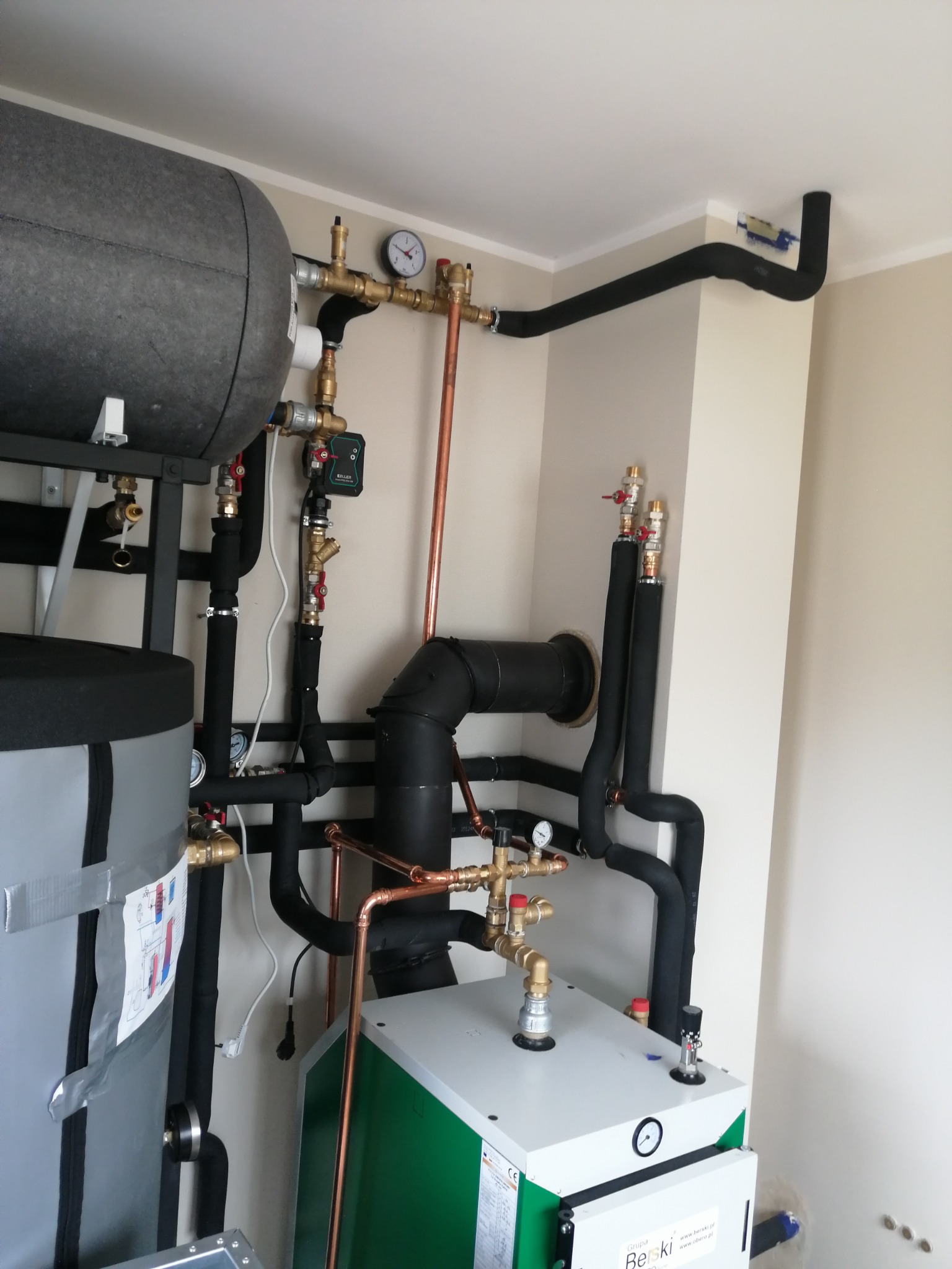 Złożona instalacja grzewcza z kotłem, zbiornikiem i rurami w izolacji, z licznymi zaworami i manometrami, na tle jasnej ściany. Widok na kotłownię.