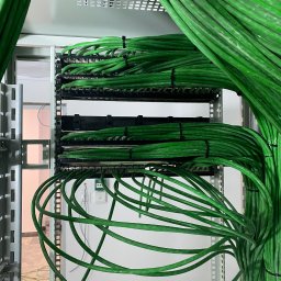 Jurkiewicz - Zorganizowane, zielone kable w szafie serwerowej, spięte czarnymi opaskami. Widoczne metalowe elementy konstrukcji i fragment pomieszczenia w tle.