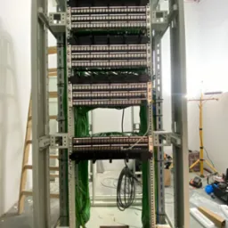 Panel krosowniczy w metalowej szafie rack z uporządkowanymi kablami. Widoczne narzędzia i drabina w tle. Instalacja teleinformatyczna w trakcie realizacji.