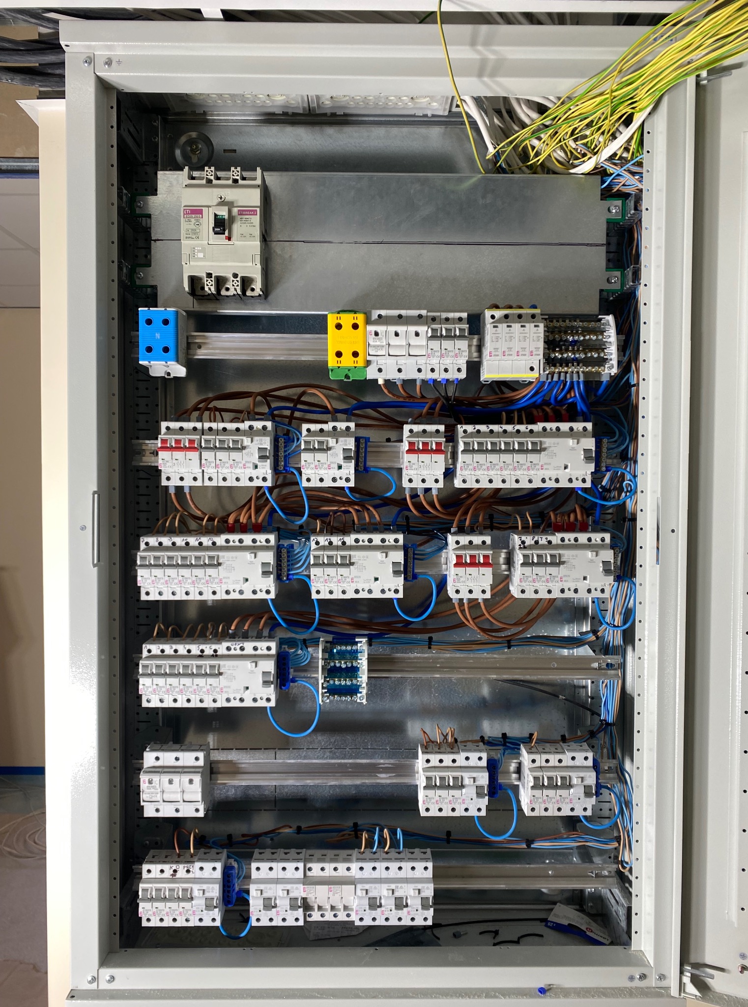 Wnętrze szafy elektrycznej z zamontowanymi bezpiecznikami, przewodami i listwami. Widoczne kable w kolorach: niebieskim, brązowym i żółto-zielonym.
