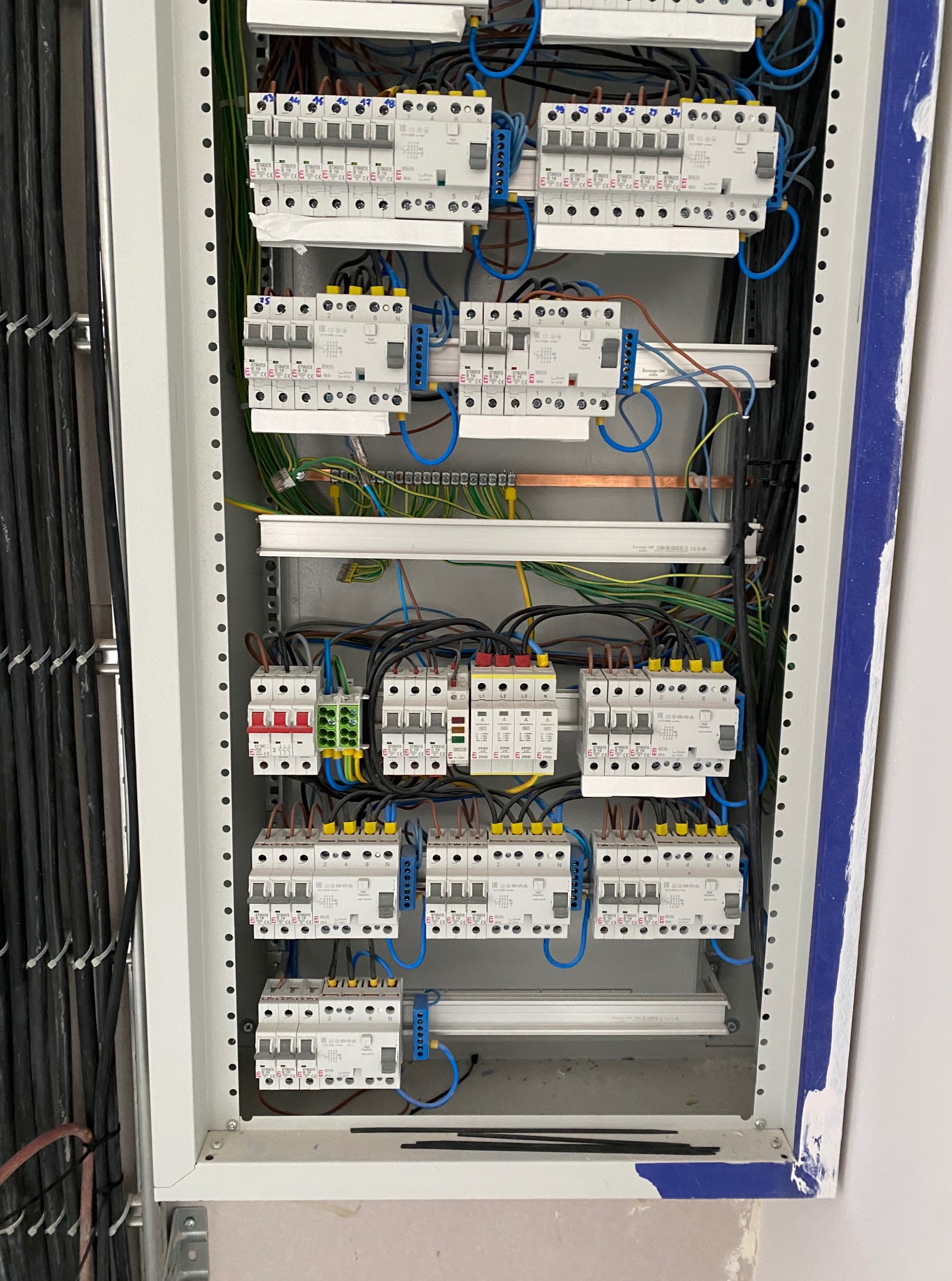 Rozdzielnia elektryczna z zamontowanymi bezpiecznikami, okablowaniem i listwami. Widoczne przewody w kolorach: niebieskim, zielonym, żółtym i brązowym.