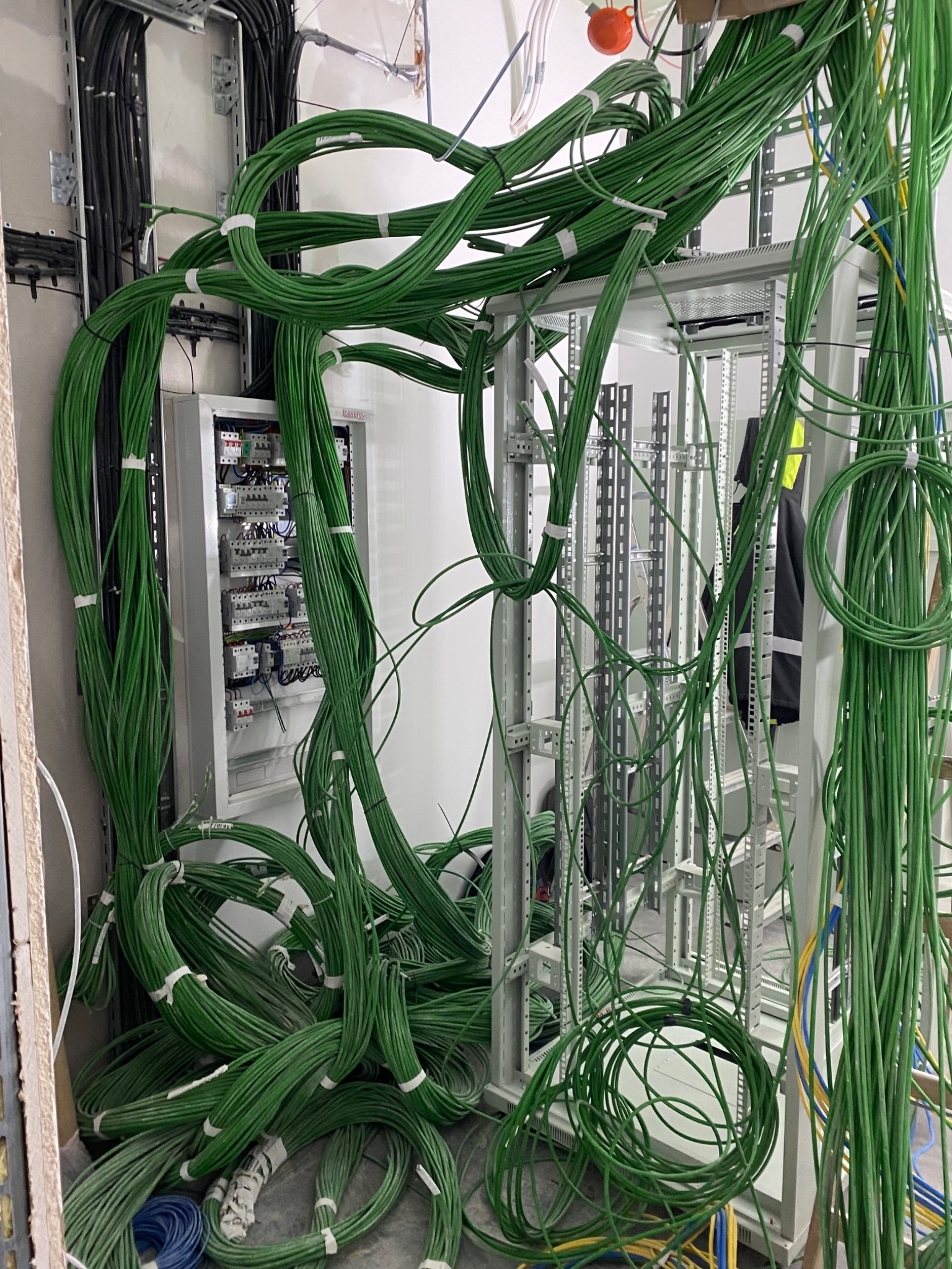 Skomplikowana instalacja elektryczna z zielonymi kablami, częściowo ułożonymi, prowadzącymi do szafy sterowniczej i tablicy rozdzielczej w surowym wnętrzu.