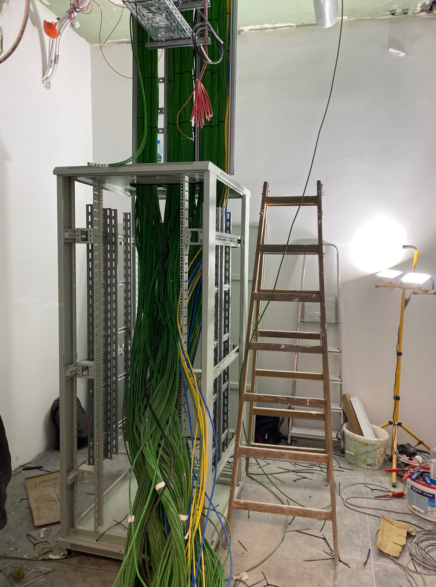 Wnętrze w trakcie instalacji elektrycznej: metalowa szafa rack z zielonymi kablami, drabina i lampa budowlana. Widoczne okablowanie pod sufitem.