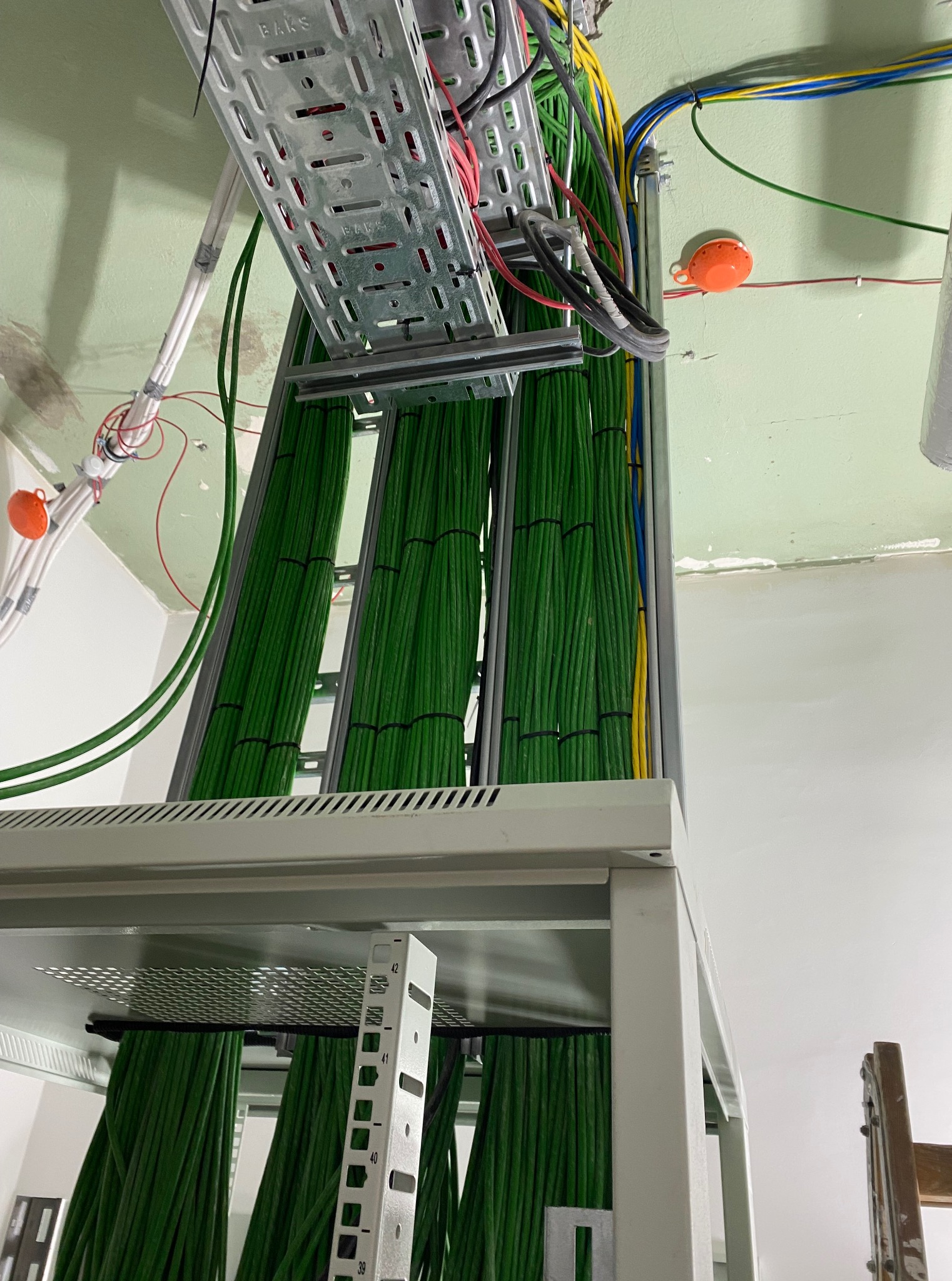 Uporządkowane kable w korytach instalacyjnych, przewody w kolorze zielonym i żółto-niebieskim, widok od dołu, szara metalowa konstrukcja wsporcza.