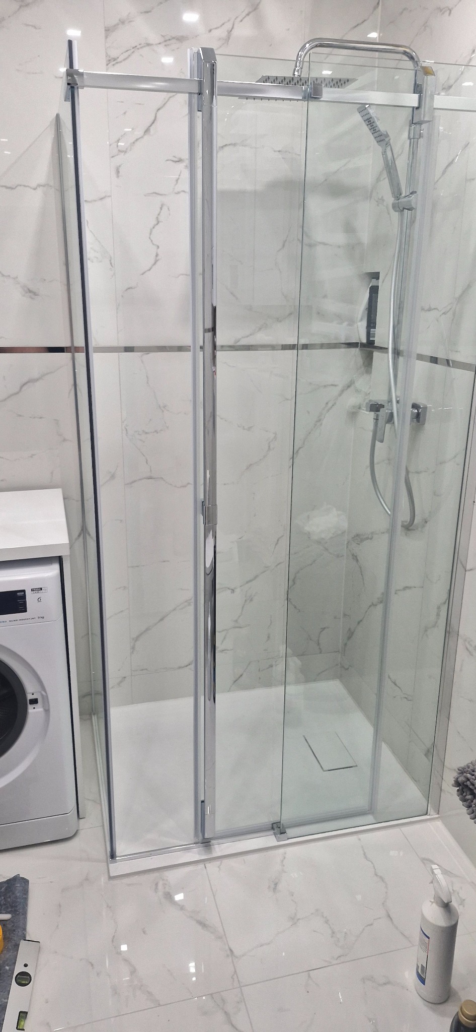 Nowoczesna kabina prysznicowa z chromowanymi profilami i deszczownicą, zamontowana w łazience wykończonej marmurowymi płytkami. Obok pralka i poziomica.