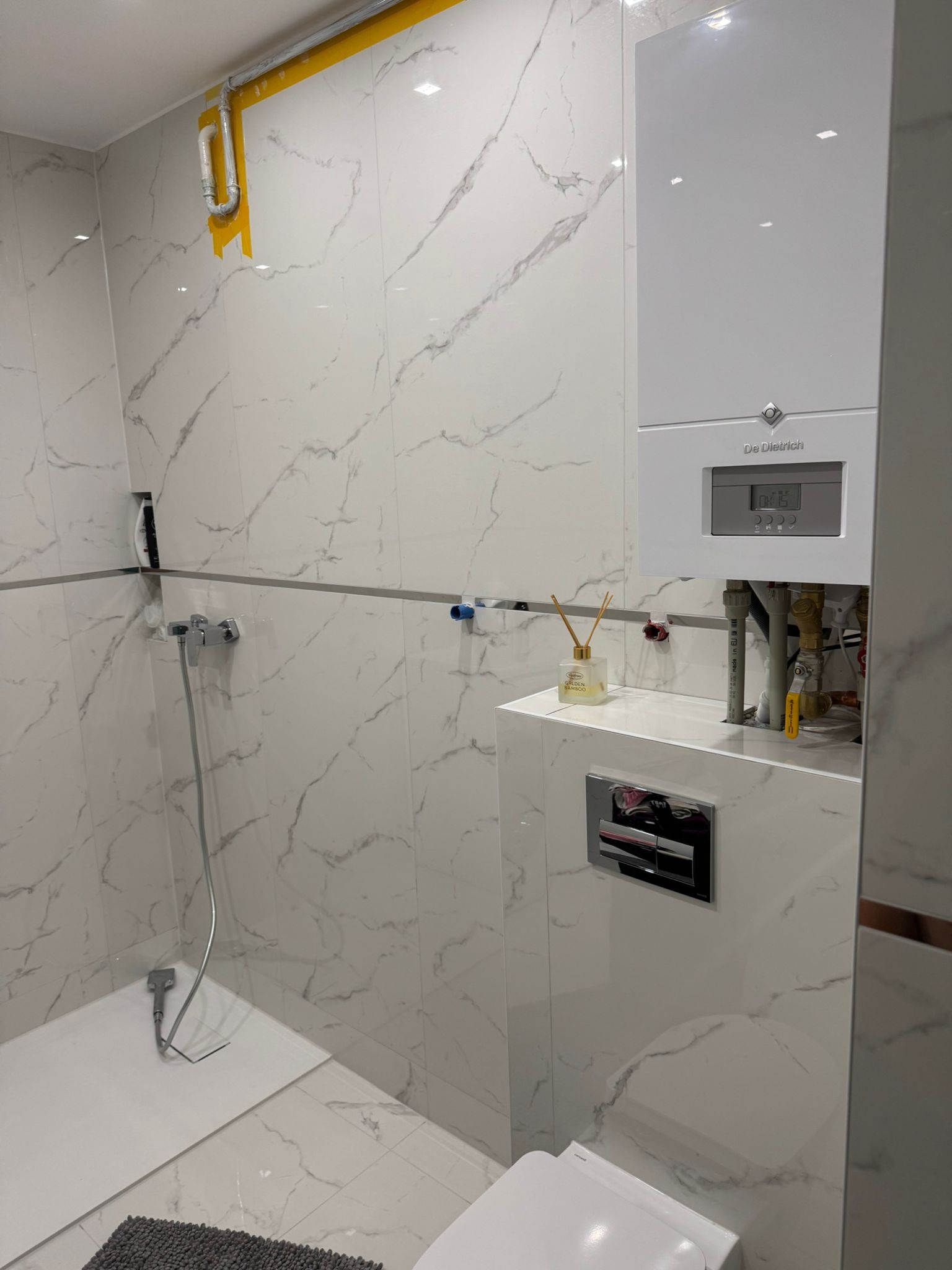 Wykończona łazienka z białymi płytkami imitującymi marmur, kabiną prysznicową i wiszącym piecem gazowym De Dietrich. Widoczne elementy instalacji i dekoracyjny dyfuzor zapachowy.