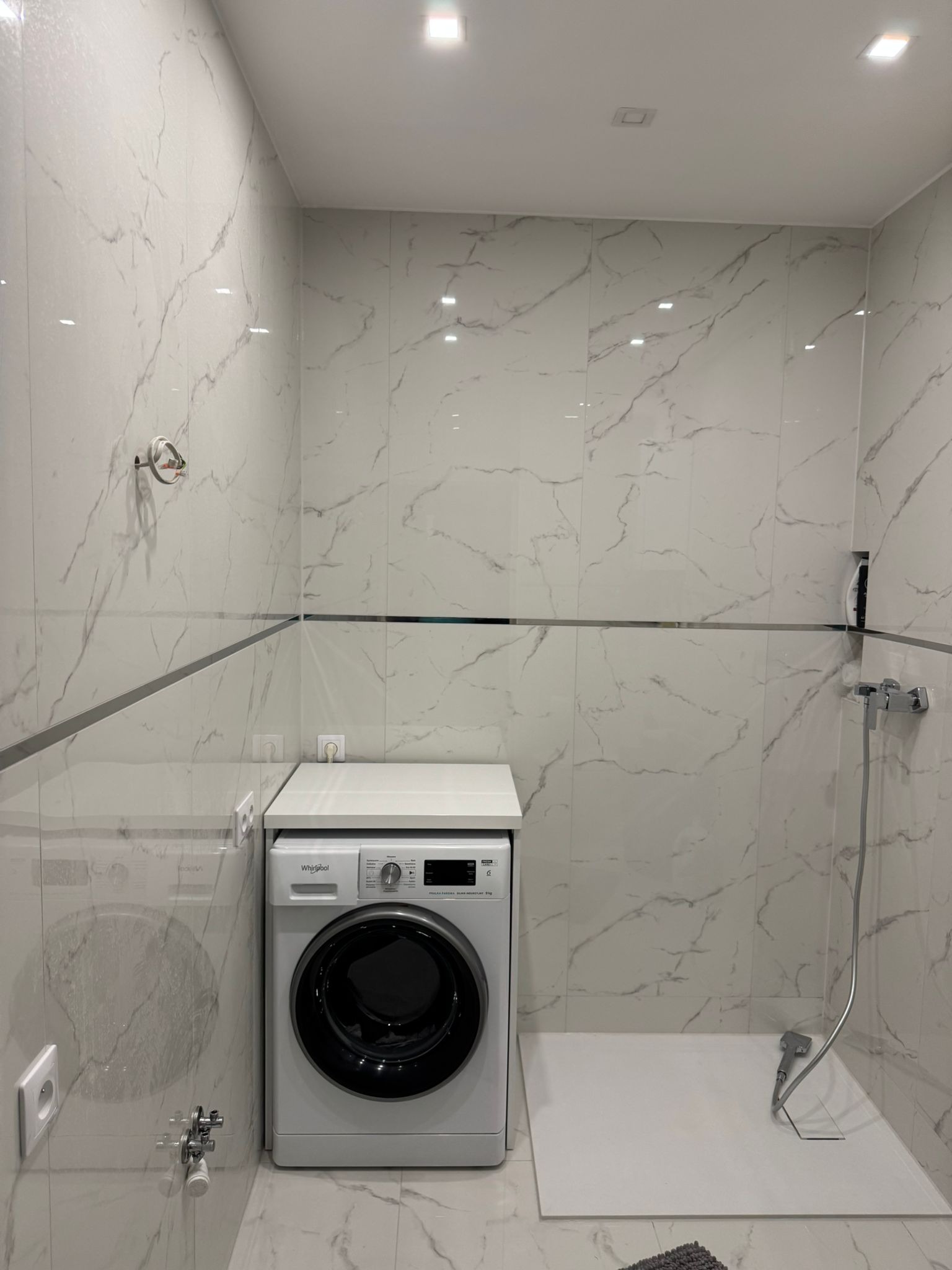 Biała łazienka z marmurowymi płytkami, kabiną prysznicową bez brodzika i pralką Whirlpool. Minimalistyczny design, chromowane wykończenia, nowoczesne oświetlenie.