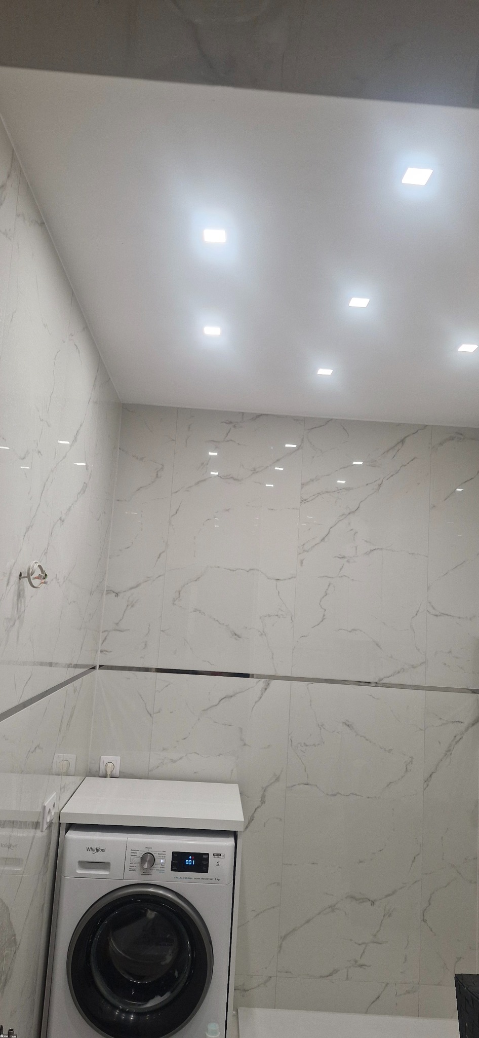 Wykończona łazienka z marmurowymi płytkami, oświetleniem punktowym i pralką. Widoczne detale wykończenia, takie jak listwa dekoracyjna. Jasne, nowoczesne wnętrze.