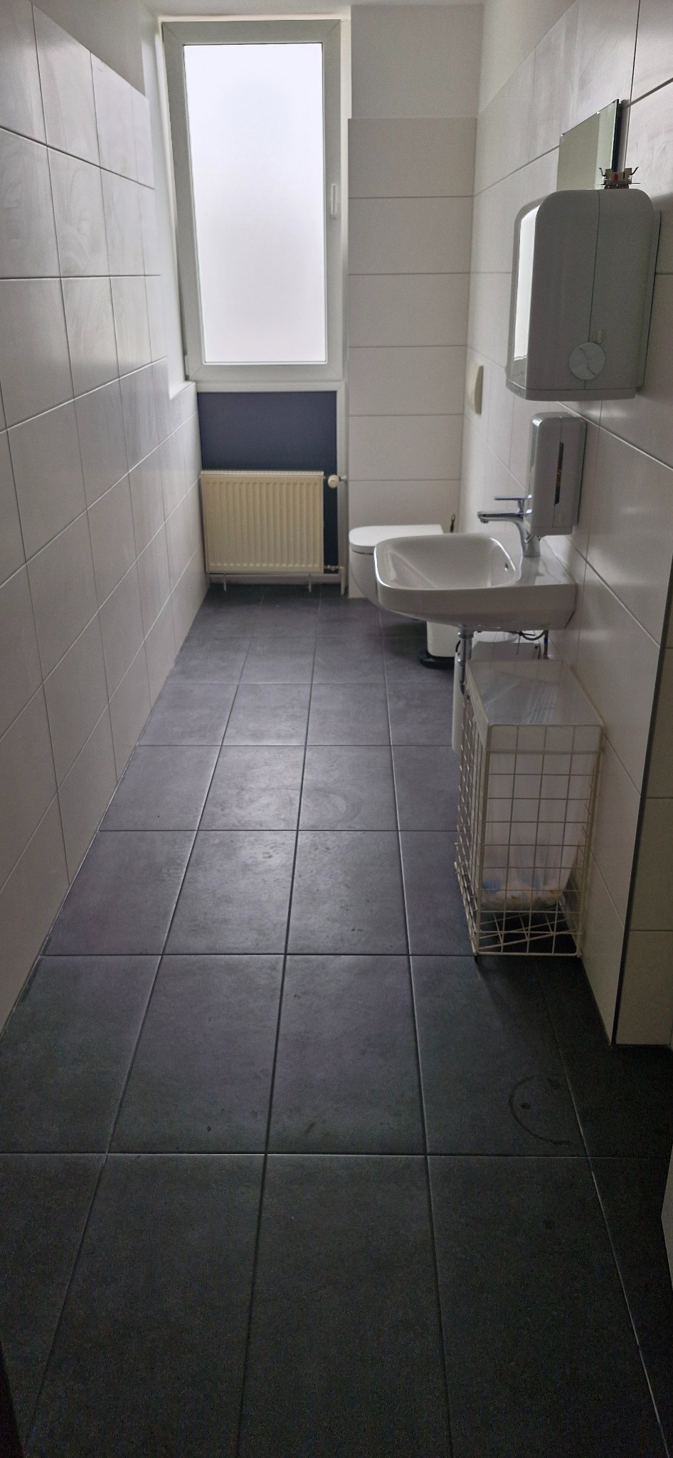 Wykończona łazienka w Radzionkowie z szarymi płytkami podłogowymi i białymi ściennymi. Widoczna umywalka, toaleta i grzejnik. Minimalistyczny design, jasne wnętrze.