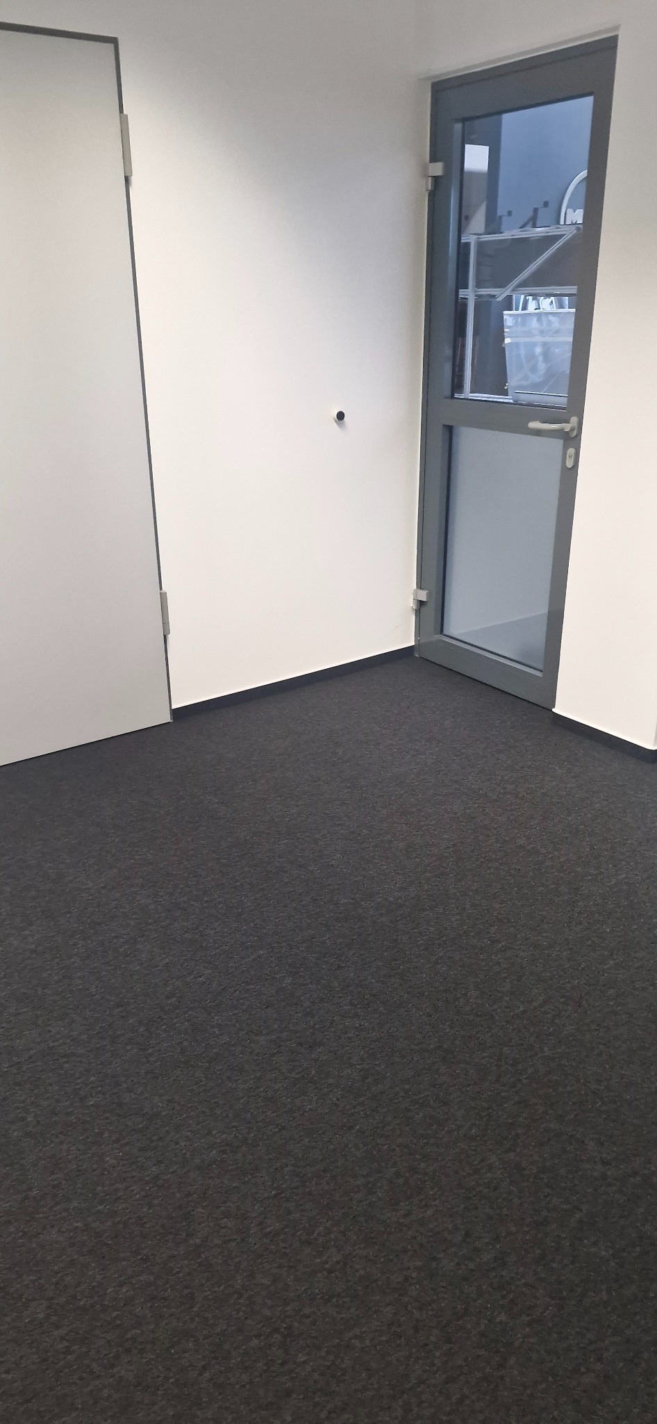 Wnętrze biura po odświeżeniu: ciemna wykładzina dywanowa, białe ściany i szare drzwi z przeszkleniami. Minimalistyczny design, czyste linie, brak dekoracji.