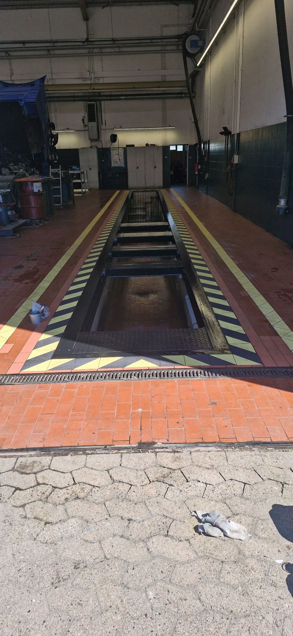 Wnętrze warsztatu samochodowego z kanałem inspekcyjnym, wyłożonym ceglaną posadzką z żółto-czarnymi pasami ostrzegawczymi. Widoczny wózek i narzędzia w tle.