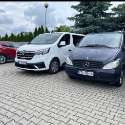 Transport osób Dominik Kubacki - Flota busów: biały Renault Trafic i granatowy Mercedes Vito, zaparkowane na kostce brukowej, z zielonymi drzewami w tle. Widoczne tablice rejestracyjne.