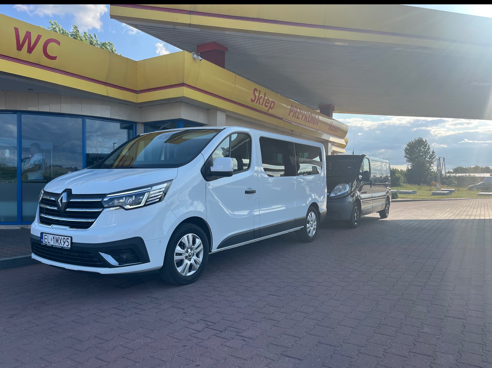 Biały bus Renault Trafic z numerem rejestracyjnym EL 1MX95 na stacji benzynowej, z widocznym drugim, ciemnym busem w tle. Słoneczny dzień.