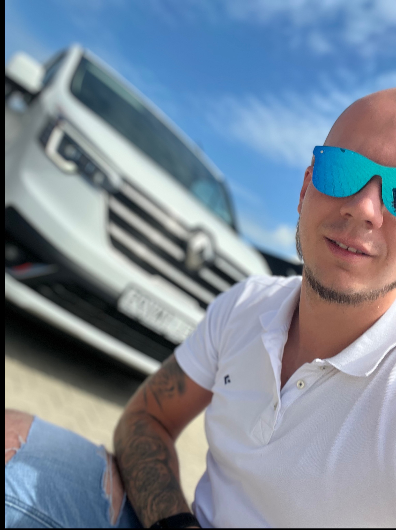 Mężczyzna w okularach z odbiciem busa w tle, na tle błękitnego nieba. Widoczne tatuaże na ramieniu i fragment podartej nogawki jeansów.