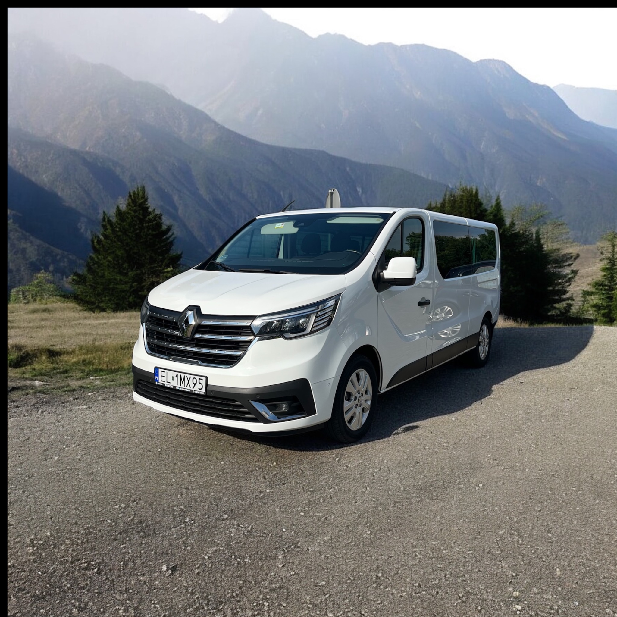 Biały bus Renault Trafic na tle górskiego krajobrazu. Pojazd zaparkowany na żwirowej drodze, widoczne drzewa i szczyty w tle. Tablica rejestracyjna EL 1MX95.