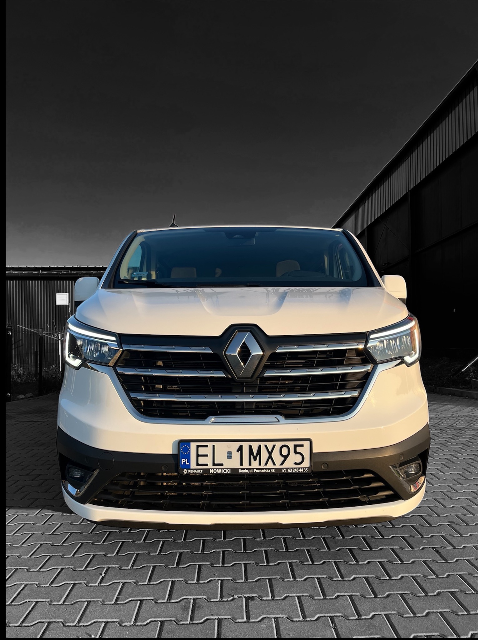 Biały Renault Trafic z tablicą EL 1MX95 na tle szarej kostki brukowej i budynku. Ujęcie z przodu, światła włączone, lekki cień. Nowoczesny wygląd.