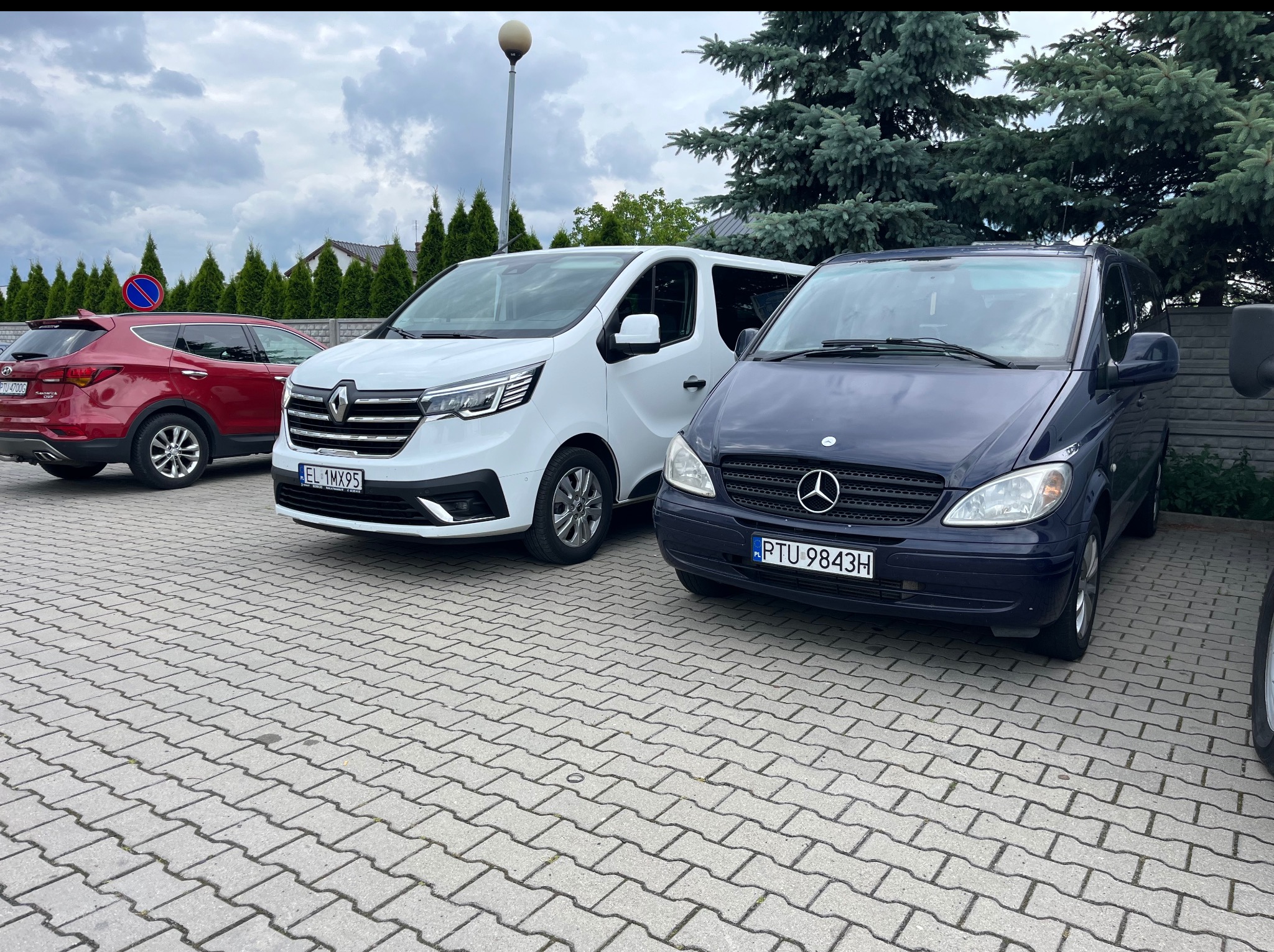 Flota busów: biały Renault Trafic i granatowy Mercedes Vito, zaparkowane na kostce brukowej, z zielonymi drzewami w tle. Widoczne tablice rejestracyjne.