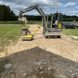 Mini koparka Volvo podnosi zagęszczarkę gruntu nad nierówną powierzchnią. W tle trawa, drzewa i czarny samochód osobowy. Niebieskie niebo z chmurami.