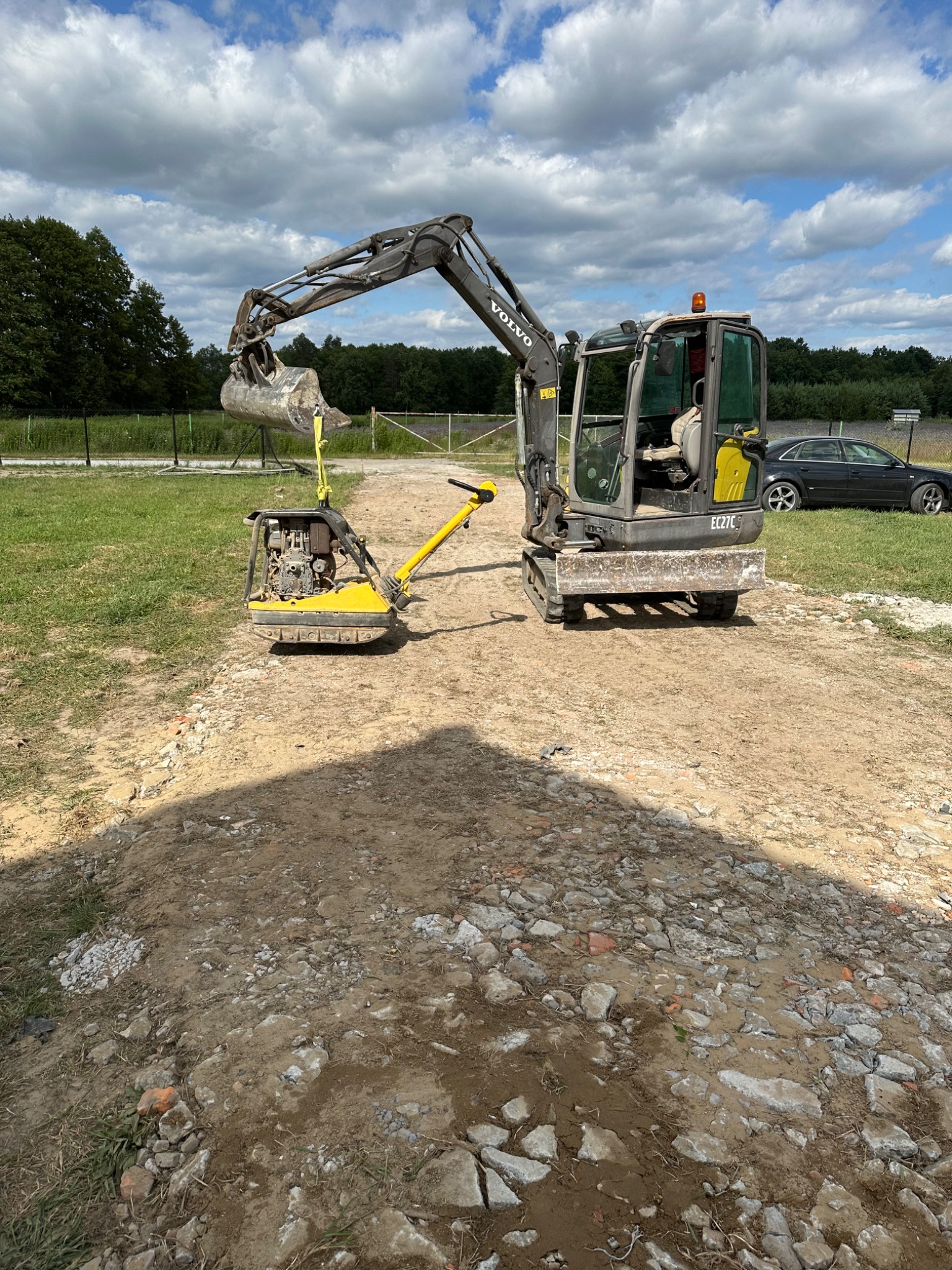 Mini koparka Volvo podnosi zagęszczarkę gruntu nad nierówną powierzchnią. W tle trawa, drzewa i czarny samochód osobowy. Niebieskie niebo z chmurami.