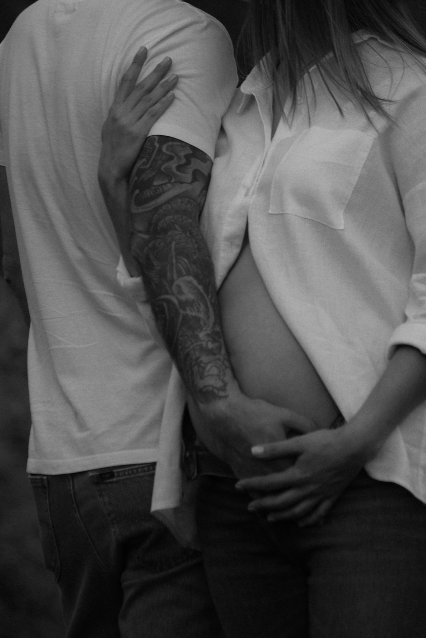Czarno-białe zbliżenie na ciążowy brzuch kobiety w rozpiętej koszuli, obejmowany przez mężczyznę z tatuażem na ramieniu. Delikatne ujęcie podkreśla więź i intymność.