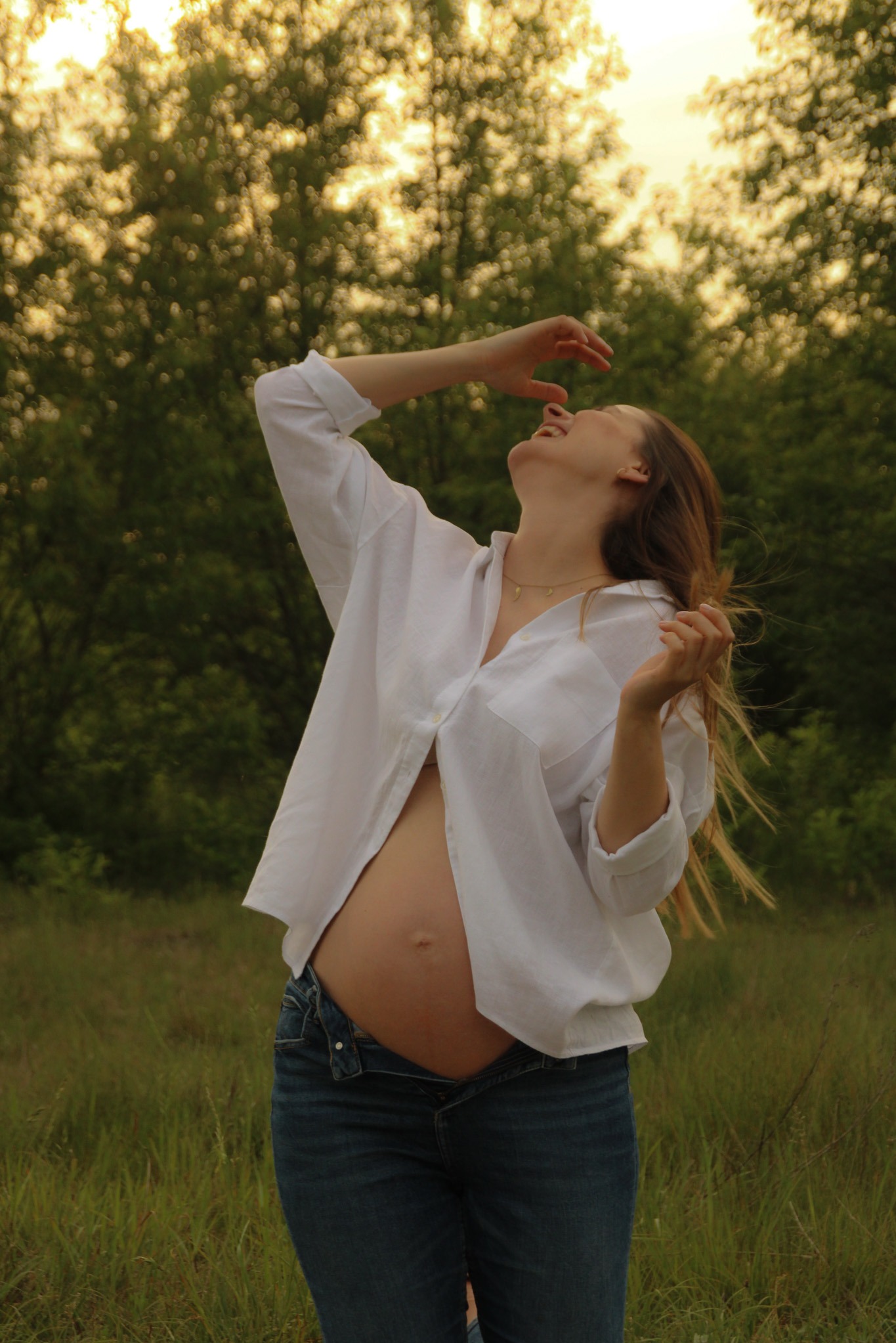 Uśmiechnięta, ciężarna kobieta w rozpiętej białej koszuli i dżinsach, z rękami uniesionymi ku górze, na tle zieleni i rozświetlonego słońcem nieba.