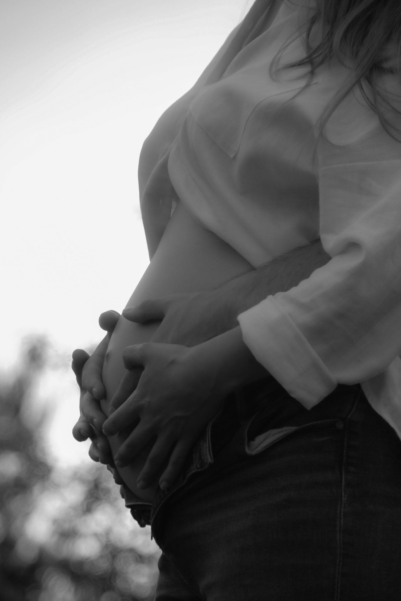 Czarno-białe zdjęcie z bliska: dłonie obejmują ciążowy brzuch kobiety w jasnej koszuli i dżinsach na tle rozmytej zieleni.