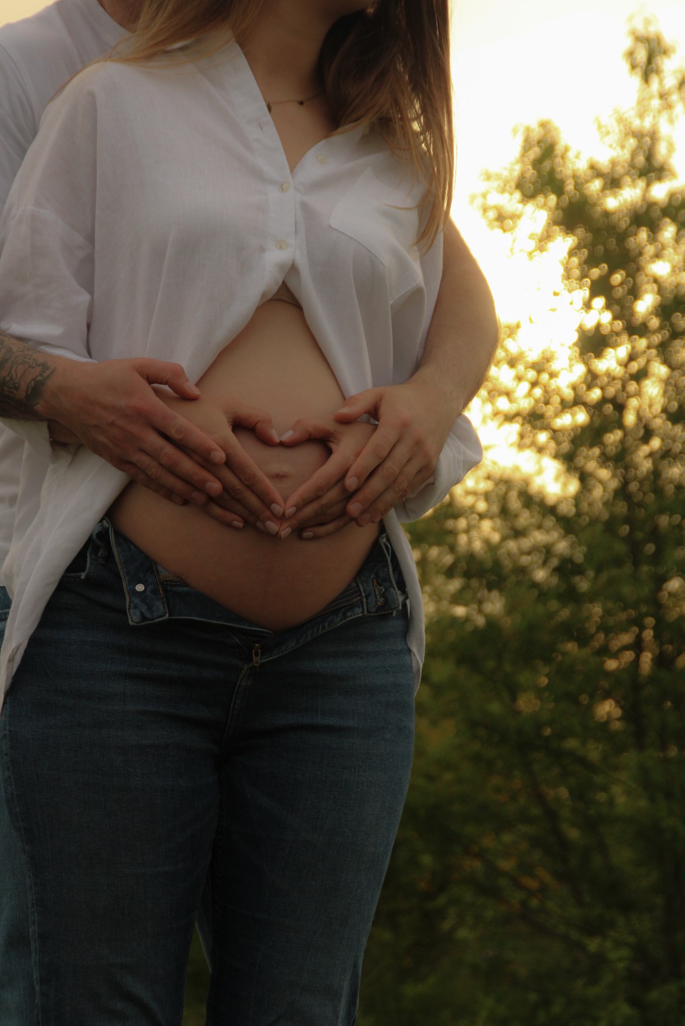 Brzuch ciężarnej kobiety, na którym dłonie mężczyzny układają się w serce. Widoczne rozpięta biała koszula i dżinsy. W tle rozmyte drzewa w ciepłym świetle.