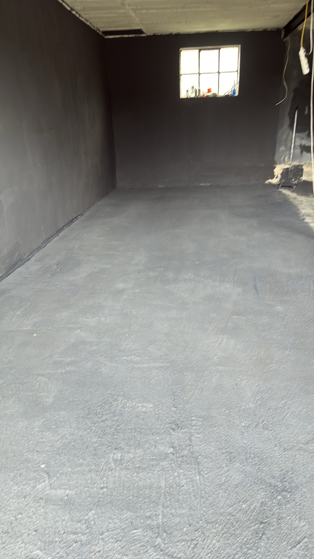 Pomieszczenie po malowaniu ścian i wylewce podłogi, widoczne okno z przedmiotami na parapecie. Szare ściany i podłoga, surowy stan.