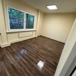 Lavrenuk Oleksandr - Jasne, puste pomieszczenie po remoncie z panelami podłogowymi. Widok na okno z białymi ramami, grzejnik i kwadratową lampę sufitową. Ściany w odcieniach beżu.