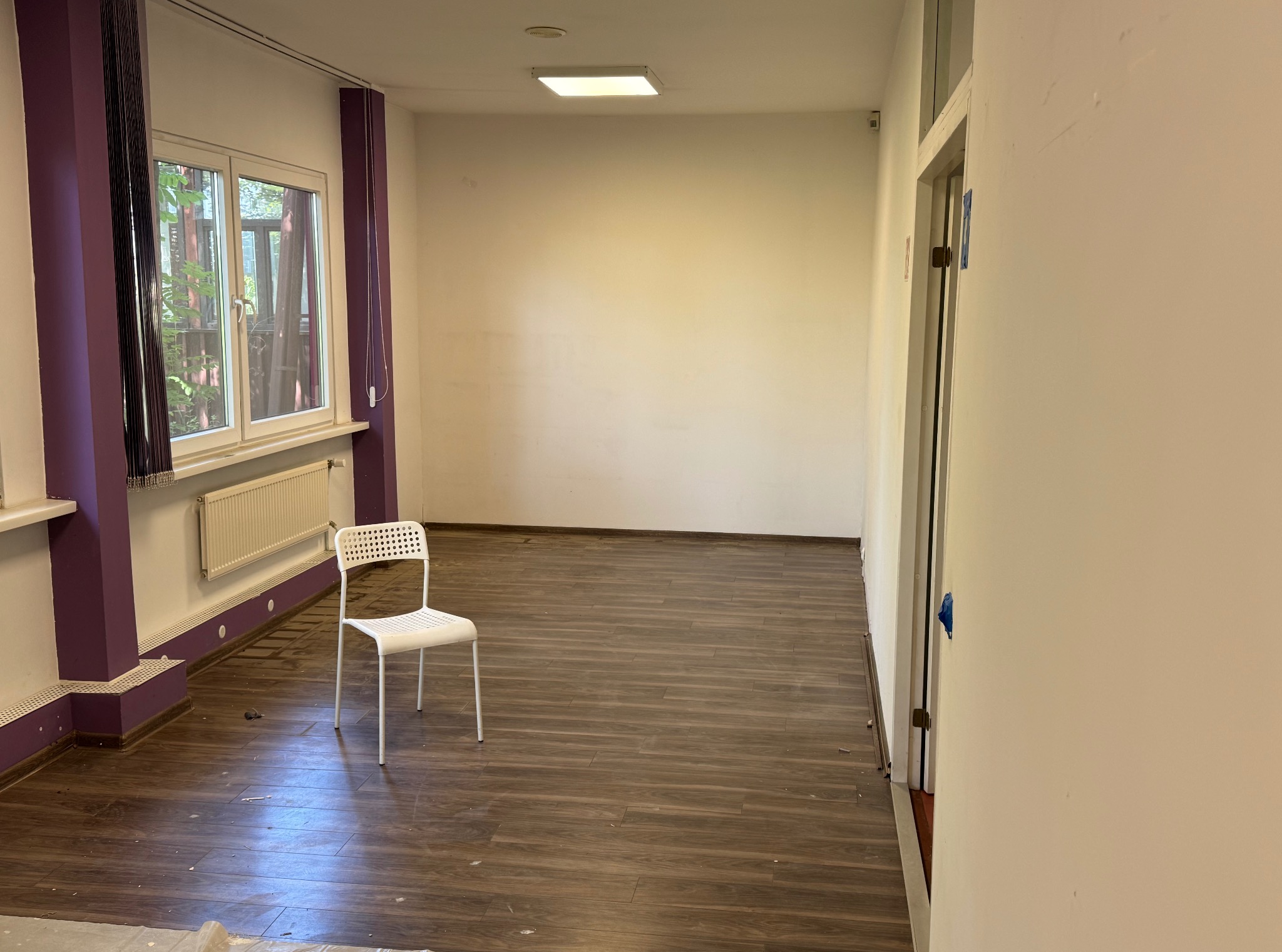 Puste pomieszczenie z brązową podłogą, białymi ścianami i fioletowymi akcentami. Białe krzesło stoi na środku. Widoczne okno z zasłonami i grzejnik.