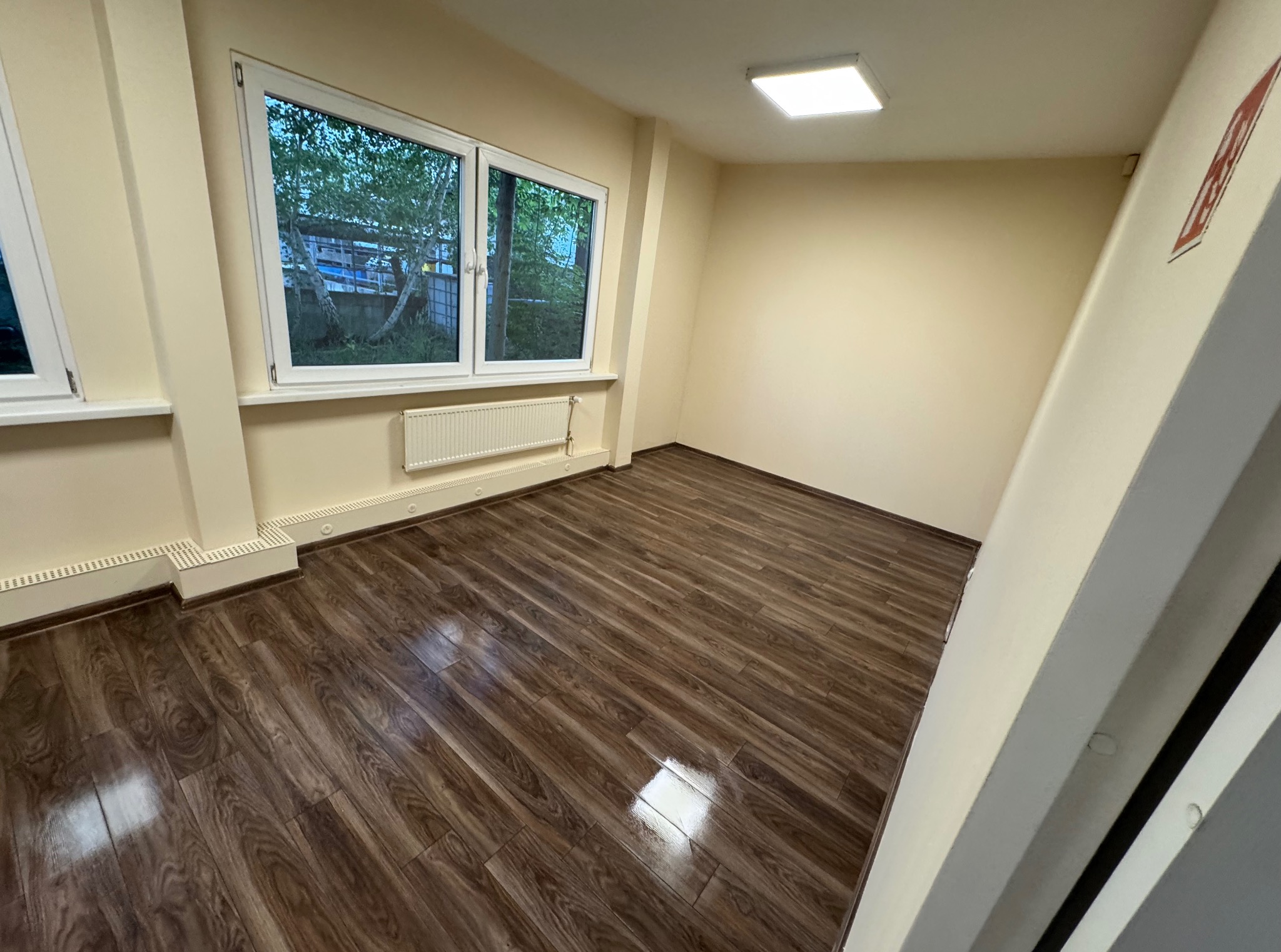 Jasne, puste pomieszczenie po remoncie z panelami podłogowymi. Widok na okno z białymi ramami, grzejnik i kwadratową lampę sufitową. Ściany w odcieniach beżu.