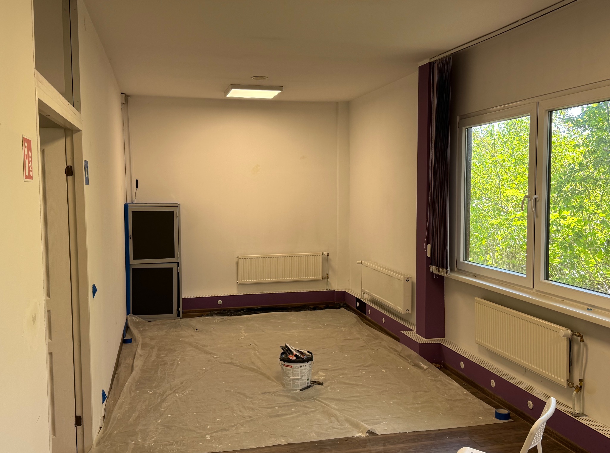 Pokój przygotowany do malowania: zabezpieczona podłoga folią, puszka z farbą na środku, pomalowane listwy przypodłogowe na fioletowo, białe ściany i grzejniki.