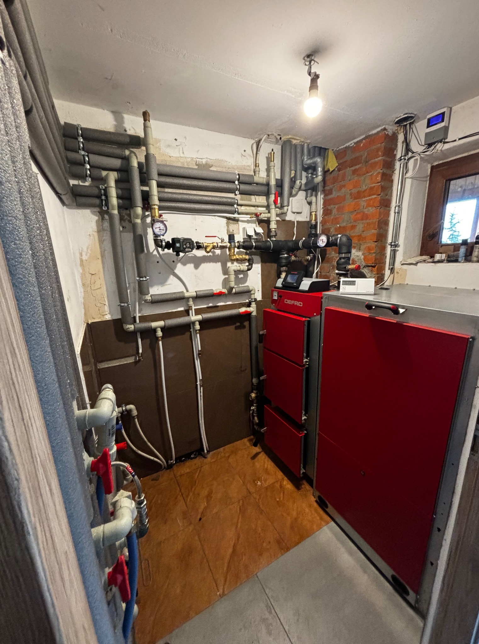 Czerwony piec Defro z instalacją w kotłowni, widoczne rury z izolacją i zawory. Ściana z cegły i okno w tle. Oświetlenie żarówką.