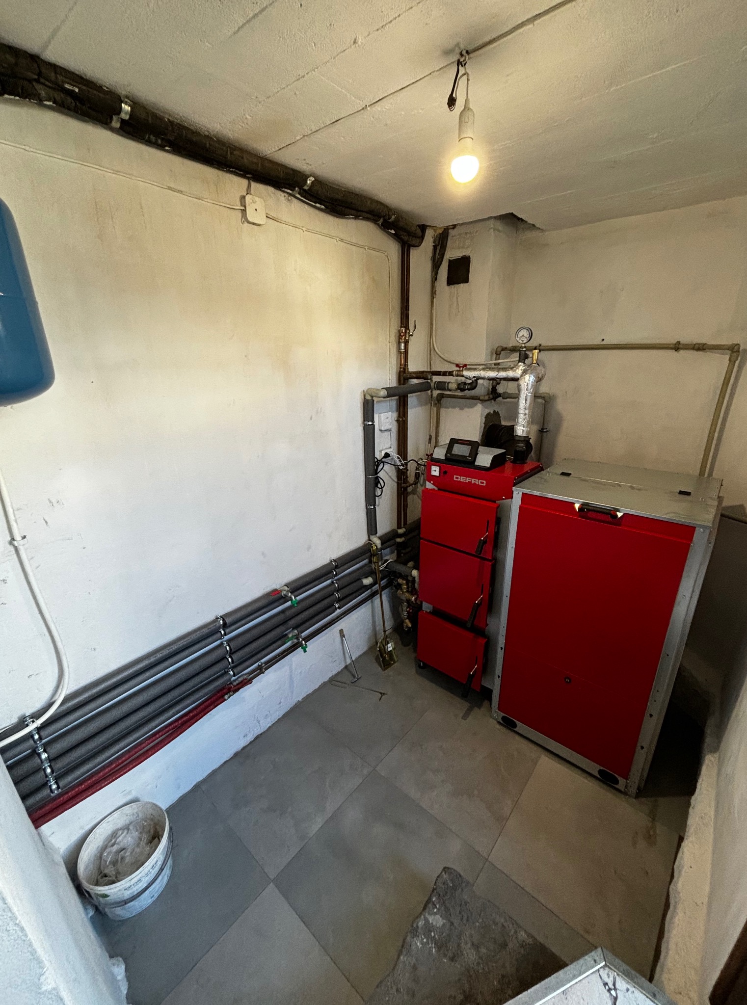 Czerwony piec Defro na paliwo stałe w kotłowni z rurami, instalacją i żarówką. Widok na wykafelkowane pomieszczenie z białymi ścianami i wiadrem.