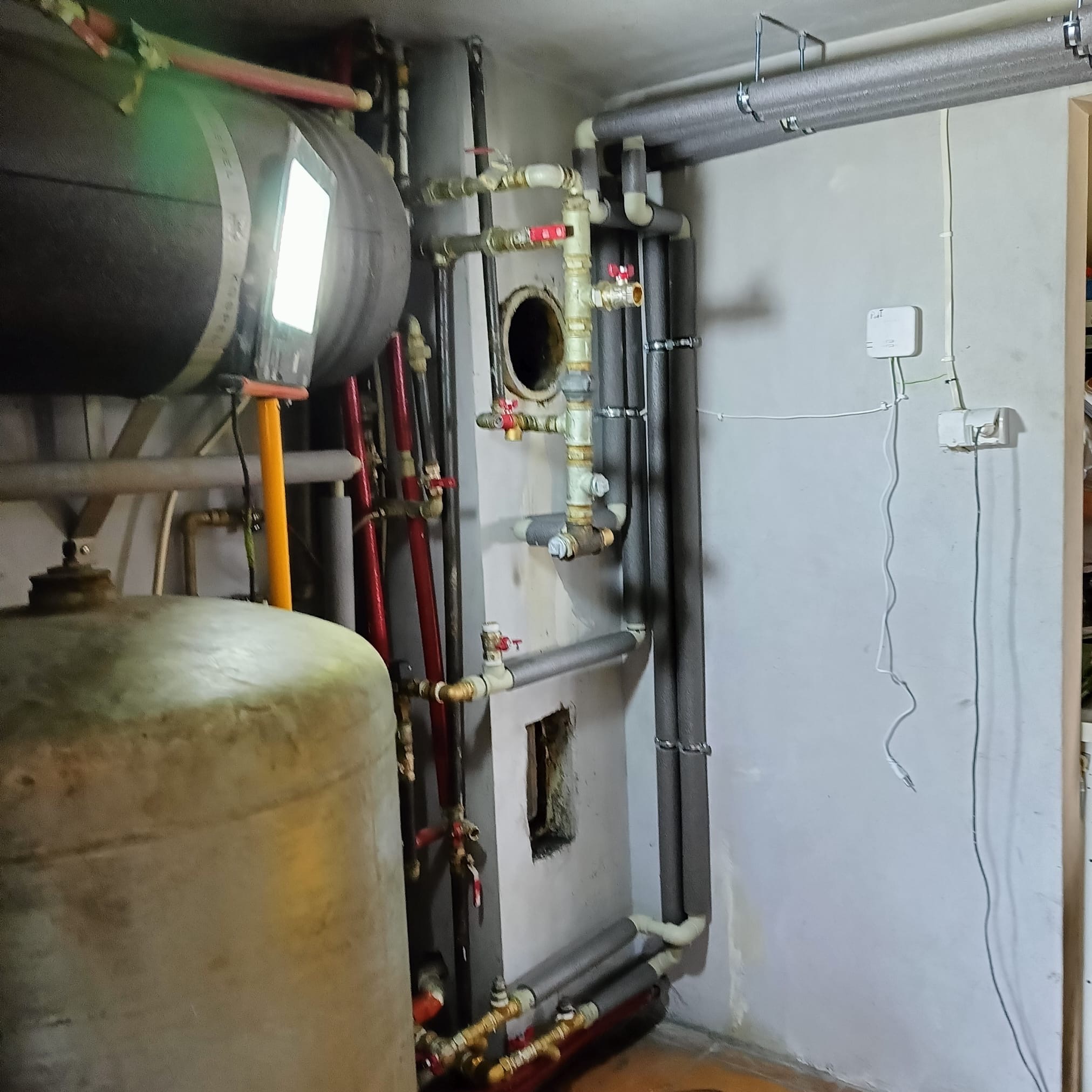 Złożona instalacja centralnego ogrzewania z zbiornikami akumulacyjnymi, rurami izolowanymi termicznie i zaworami. Widoczne elementy sterujące i okablowanie.
