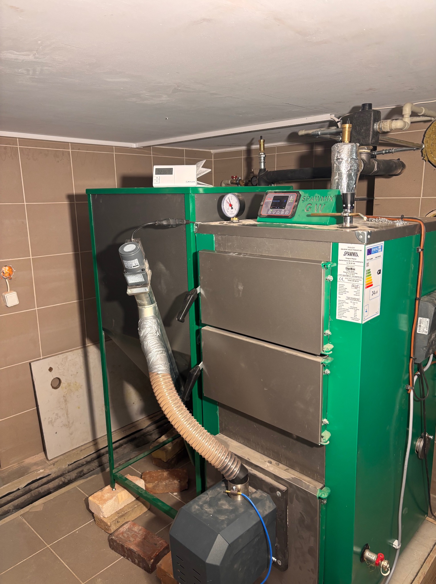 Zielony piec na paliwo stałe marki Spawmix w kotłowni, z widocznymi elementami instalacji i cegłami podtrzymującymi konstrukcję. Ujęcie z bliska, z widocznymi detalami.