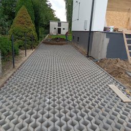 RYSZARD CHRZANOWSKI - Ułożona szara kostka ażurowa na podjeździe, widoczny fragment budynku i prace ziemne w tle. Kostka ułożona równo, tworząca wzór. Prace brukarskie w toku.