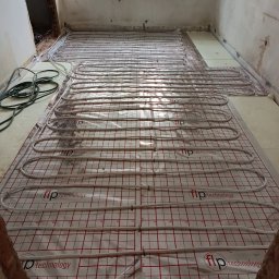 Usługi ABC - Instalacja ogrzewania podłogowego z białych rur na folii z czerwoną kratką w surowym wnętrzu remontowanego pomieszczenia. Widoczne kable i okno w tle.