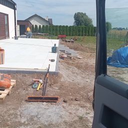 RYSZARD CHRZANOWSKI - Jasnoszara kostka brukowa na tarasie w trakcie układania. Widoczne narzędzia, materiały budowlane i betoniarka. W tle dom z brązowymi elementami.