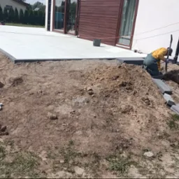 Montaż tarasu z jasnych płyt na gruncie. Dwóch pracowników układa obrzeża z szarej kostki. Widoczny paleta z narzędziami i materiały budowlane.