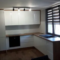 Nowoczesna kuchnia z białymi, lakierowanymi frontami i drewnianymi blatami. Minimalistyczne oświetlenie punktowe i okno z żaluzjami. Widok na zabudowę kuchenną.