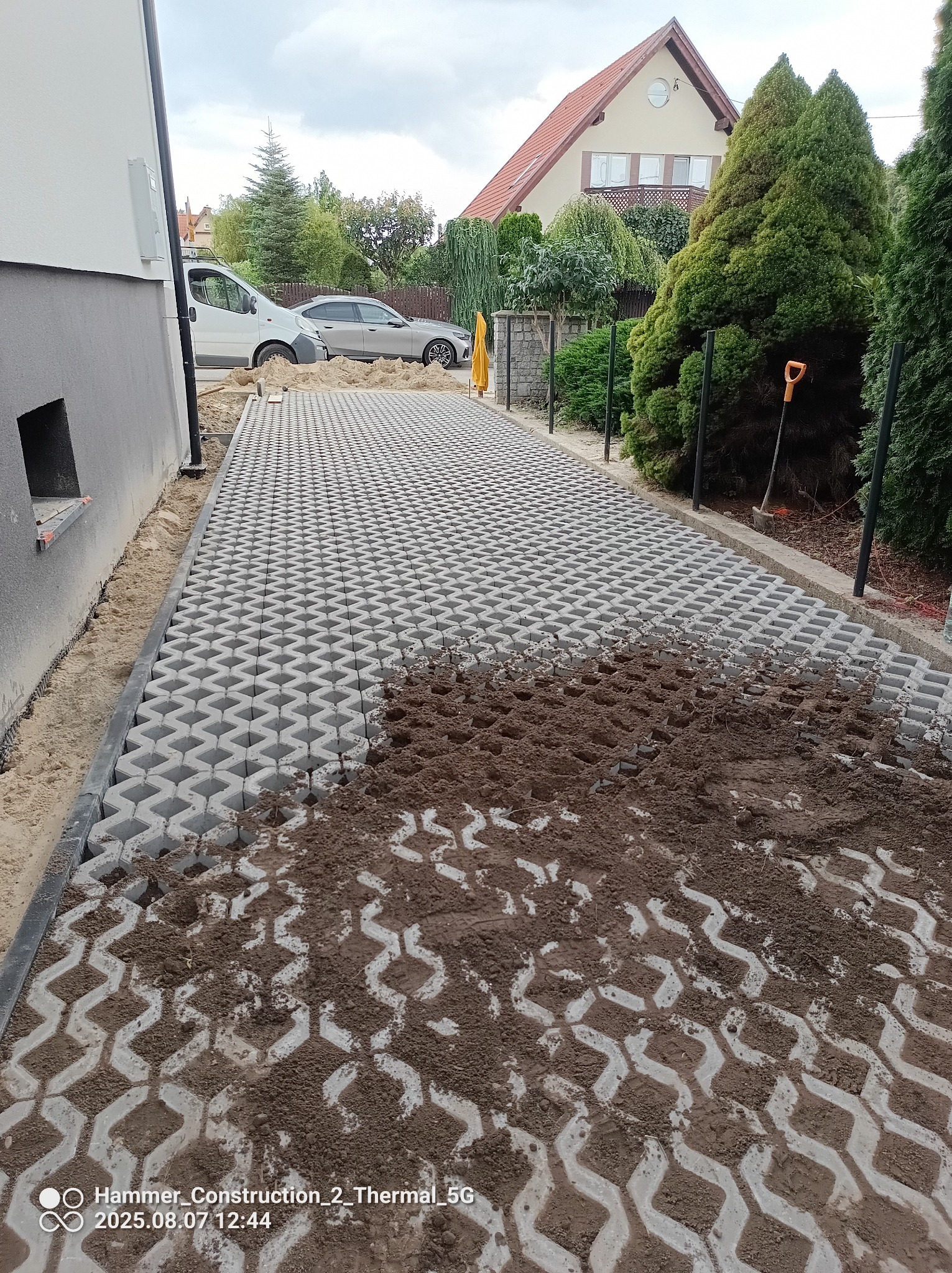 Częściowo wypełniona ziemią, szara ażurowa kostka brukowa na posesji z widokiem na budynek mieszkalny i samochody. Etap prac brukarskich.